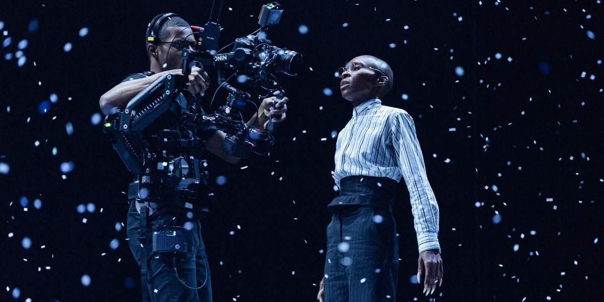 Production shots for Bram Stoker's Dracula starring Cynthia Erivo. She wears a white shirt and glasses, gazing into a camera held by a cameraman. She is surrounded by white confetti falling around her.