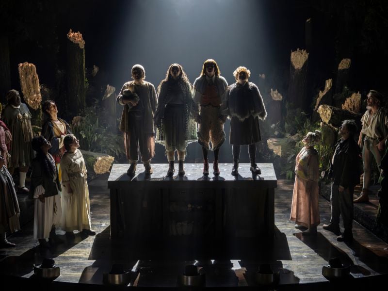 Into The Woods production shot at the Bridge Theatre. Four cast members stand on a block with a bright white light behind them, creating a shadowy silhouette. The ensemble all look up at them surrounding the block. 