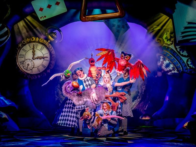 Production shot from Alice in Wonderland at Riverside Studios. Alice is surrounded by the characters of Wonderland, including an owl and a Dodo bird.