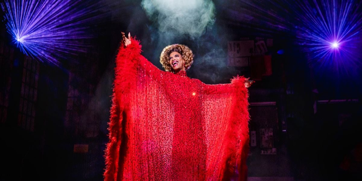 Johannes Radebe as Lola, dressed in a sequined red mesh dress with the lining decorated with red feathers, singing with their arms spread open towards the audience (Photography by Matt Crockett).