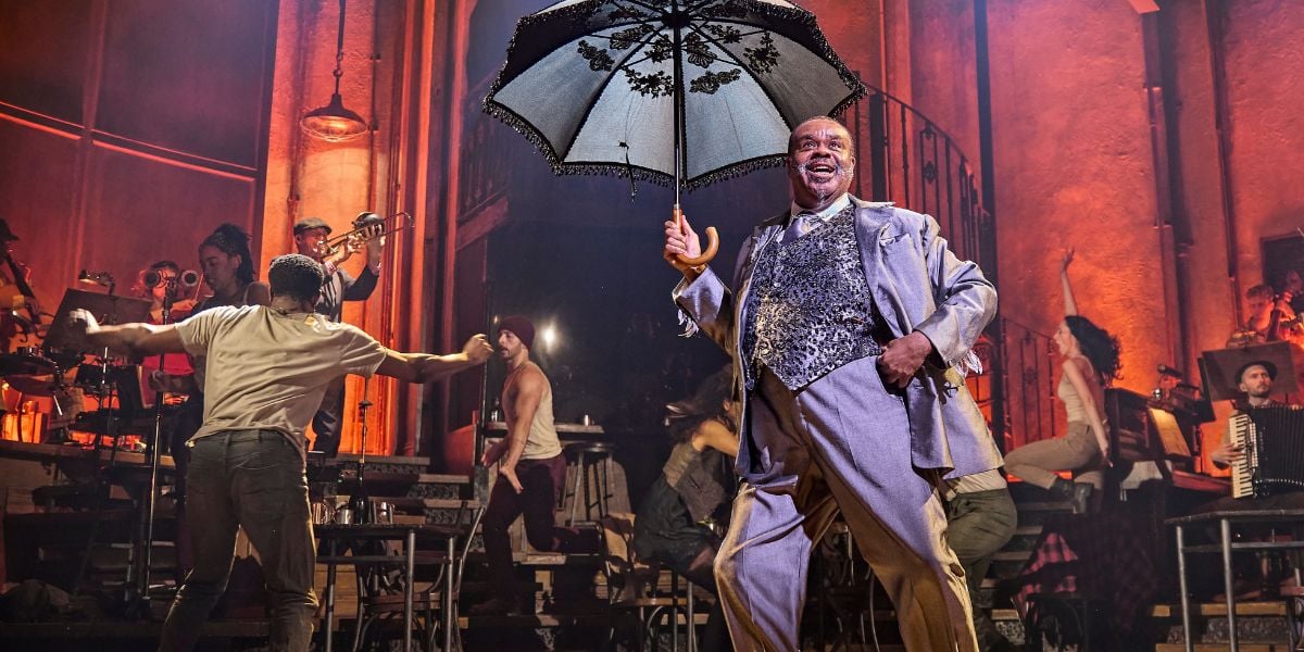 Clive Rowe in a grey suit holding an umbrella in Hadestown. © Brinkhoff-Moegenburg