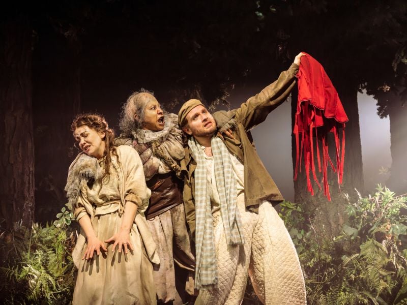Into The Woods production shot at the Bridge Theatre. From left to right: Rachel Tucker (Baker’s Wife), Melanie La Barrie (Witch) and Hughie O’Donnell (the Baker), he holds a red cloak in the air whilst the witch shouts at him and clutches them both.