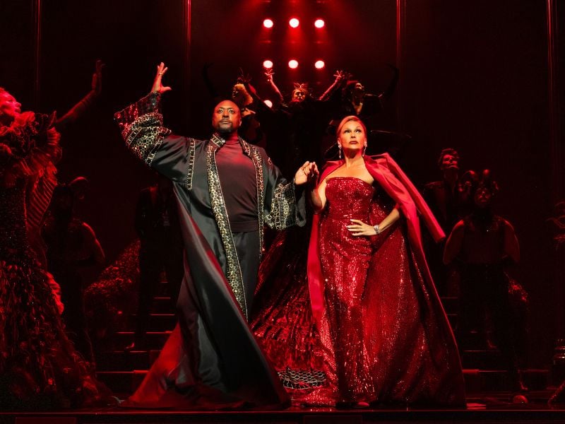 Vanessa Williams as Miranda Priestly in The Devil Wears Prada Musical in London. She wears a red floor length gown and a red cape. Nigel by her side holds her hand.