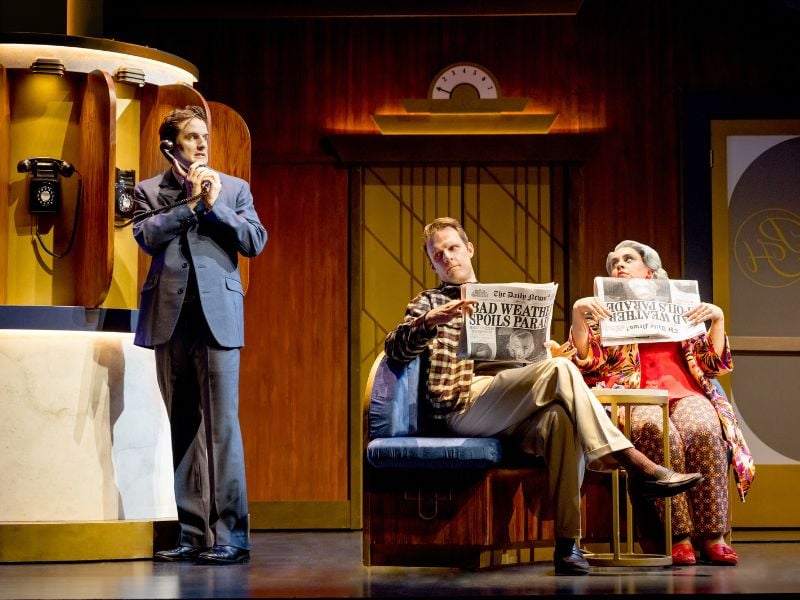 Production shot from The Comedy About Spies. One man in a suit looks nervous, looking over his shoulder on the phone, while two others listen in from behind their newspapers.
