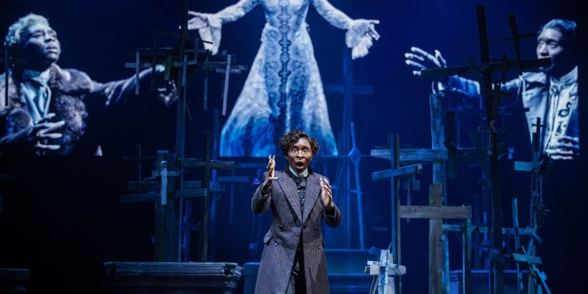 Production shots for Bram Stoker's Dracula starring Cynthia Erivo. She wears a trench coat with a short haired wig and glasses talking out to the audience with her hands.
