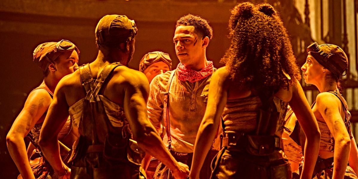 Marley Fenton & the cast of Hadestown. In a dim warm lighting, Fenton is surrounded by the cast with blood on his face. © Brinkhoff-Moegenburg