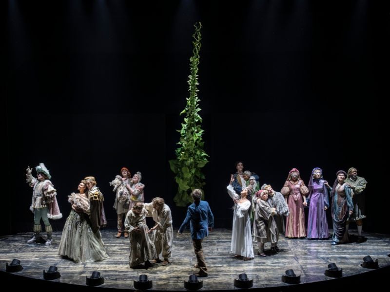 Into The Woods production shot at the Bridge Theatre. A large beanstalk ascends from center stage. The ensemble look into the air, whilst Jo Foster (Jack) looks up at the beanstalk.