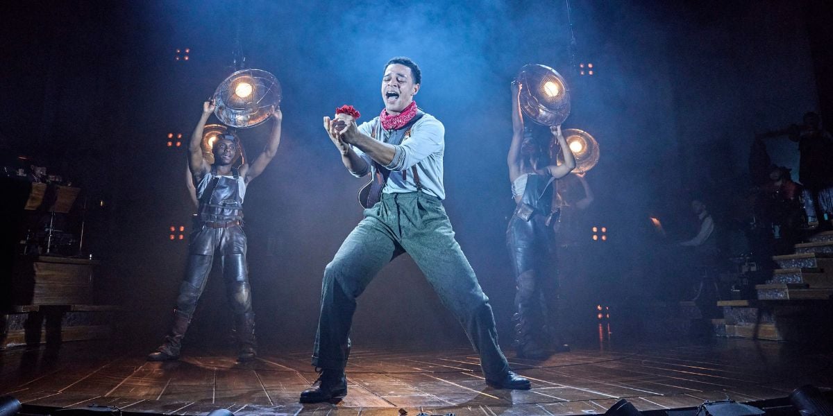 Marley Fenton & the cast of Hadestown. He holds a red flower in his hands passionately singing. © Brinkhoff-Moegenburg