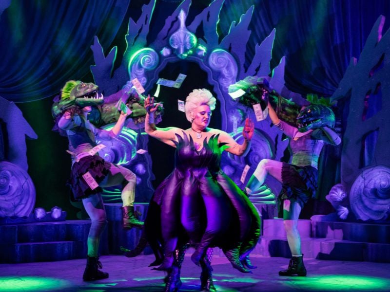 A production shot from Unfortunate. Ursula the sea witch is stood between two sea creatures who are raining money on her.
