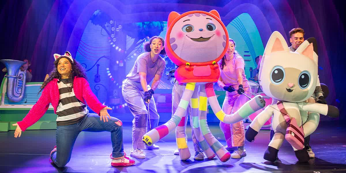 Gabby kneels with her hands out towards the audience. There are two large cat puppets also facing the audience.