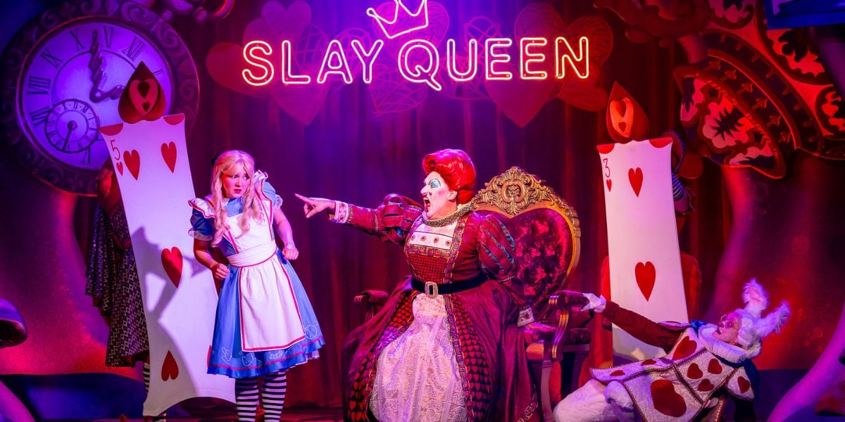 Alice in Wonderland at Riverside Studios. The Queen of Hearts (Daniel Page) is pointing at Alice (Charlotte Bradley) screaming at her with rage. Behind them are two heart playing cards and a neon sign saying 'slay queen.'