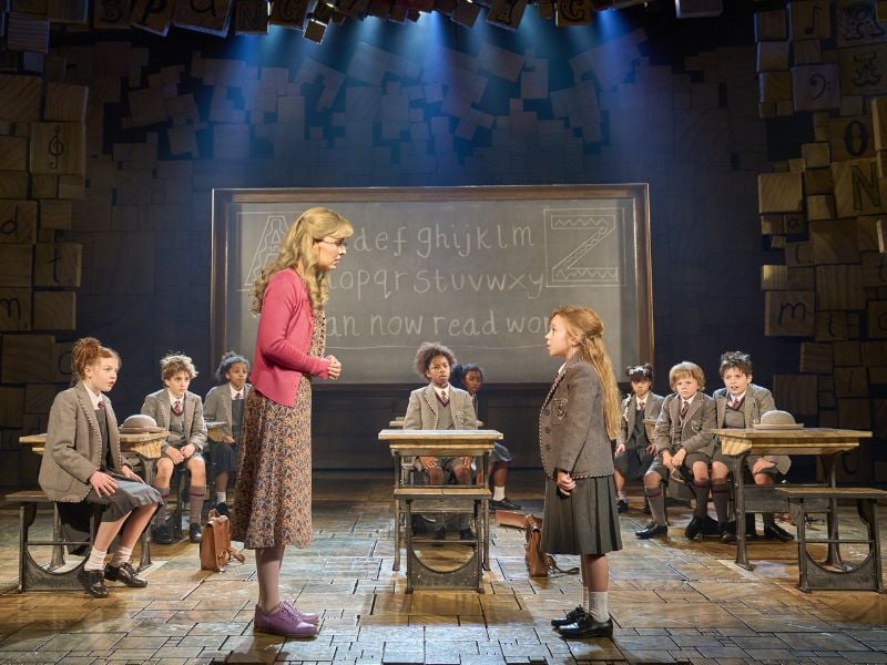 Production photo from Matilda the Musical. Matilda wears a grey school uniform and is looking up at Miss Honey while the class of children watch their exchange. 