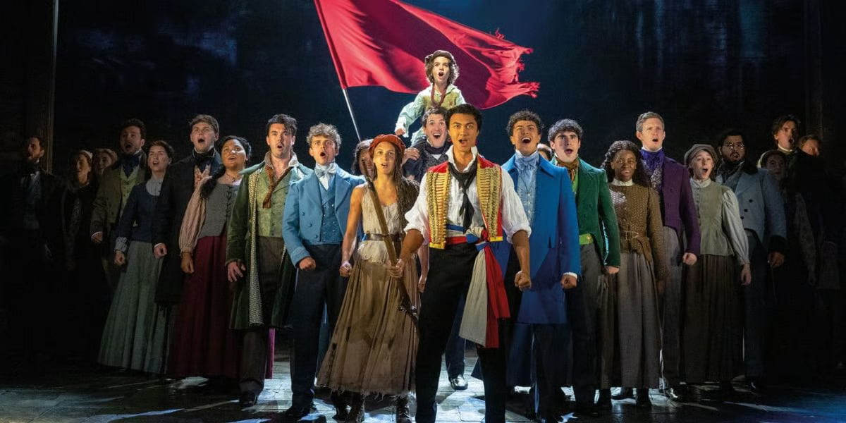 Les Misérables production image of the barricade scene showing revolutionaries holding flags and rifles in a dramatic stage setting from the West End musical.