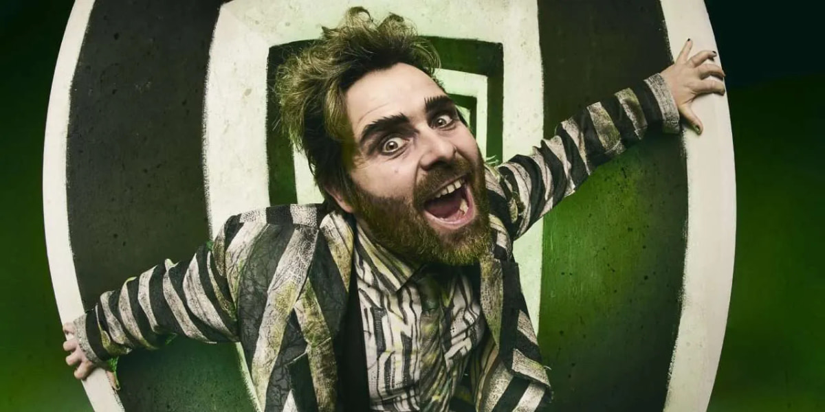 Beetlejuice the Musical star David Fynn, dressed in the iconic black-and-white striped Beetlejuice suit, leans wildly toward the camera with a manic, comic expression, capturing the chaotic, high-energy tone of the production.