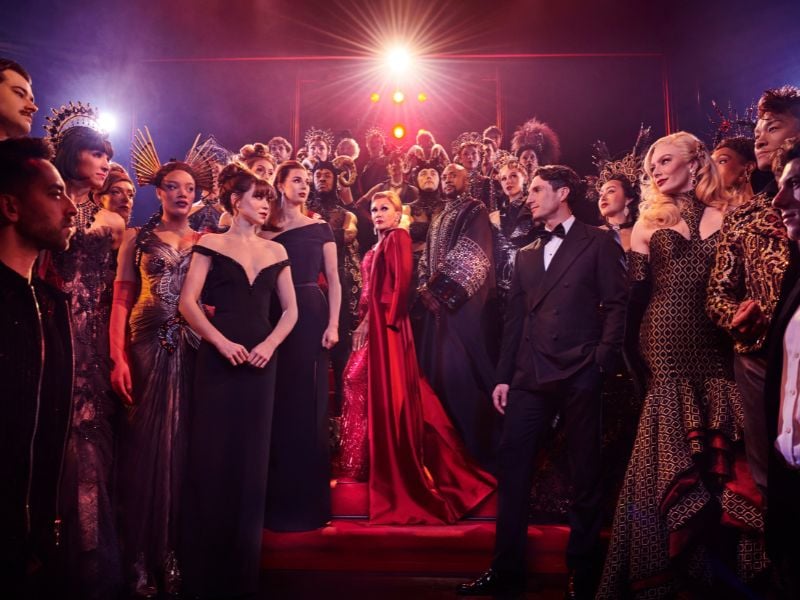 Production shot of The Devil Wears Prada musical cast. Vanessa Williams as Miranda Priestly stands centre stage, all in red whilst the cast look in awe at her.