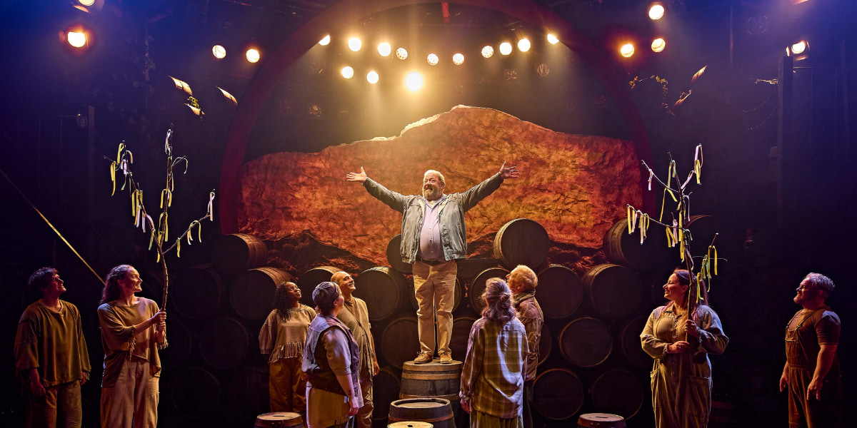 Man stands on a barrel with arms raised, surrounded by ensemble cast in a warm stage scene from Harold Fry the musical