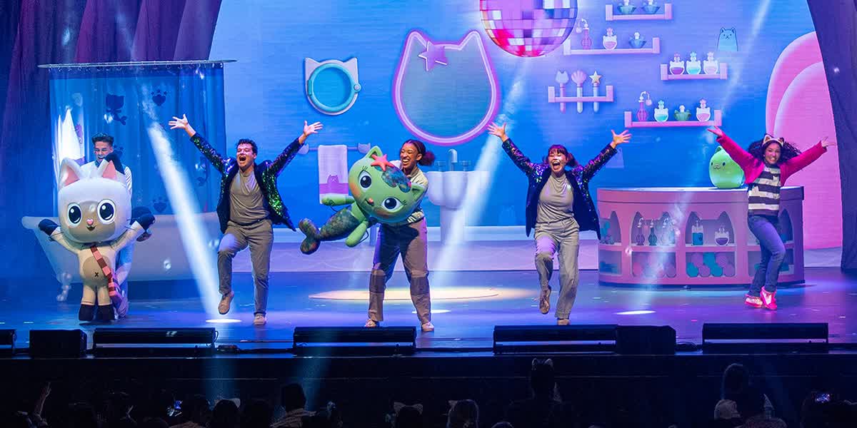 Two cat puppets, two performers and the main character Gabby hold their hands in the air on stage.