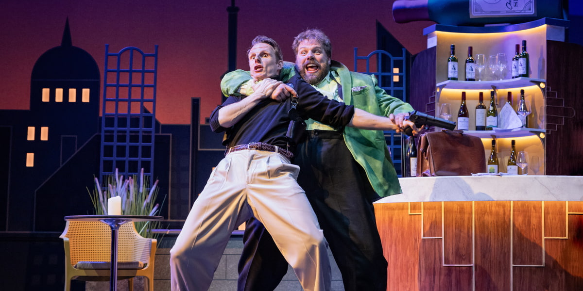 Two actors in The Comedy About Spies perform a physical comedy moment on stage beside a bar set, with one actor holding the other in an exaggerated headlock against a stylised city skyline backdrop.