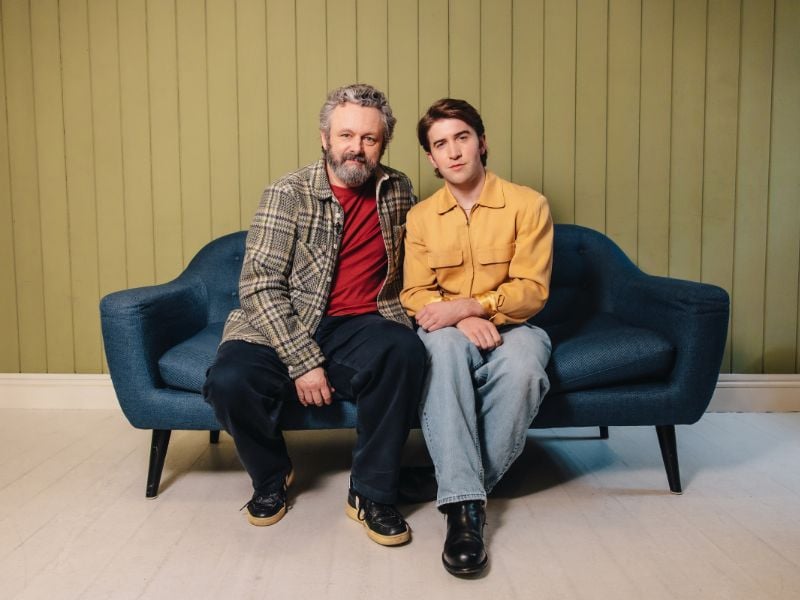 Michael Sheen and co-star Callum Scott Howells sitting on a sofa posing for the camera.