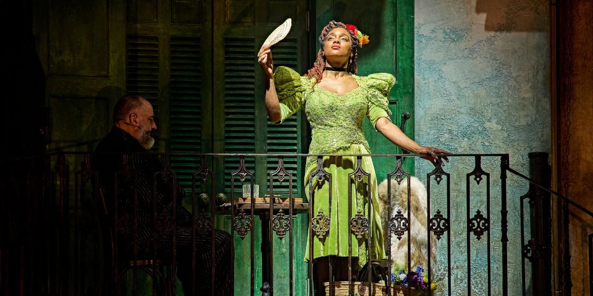 Rachel Adedeji wears a green dress on a balcony with a fan in her hand looking up in Hadestown. © Brinkhoff-Moegenburg