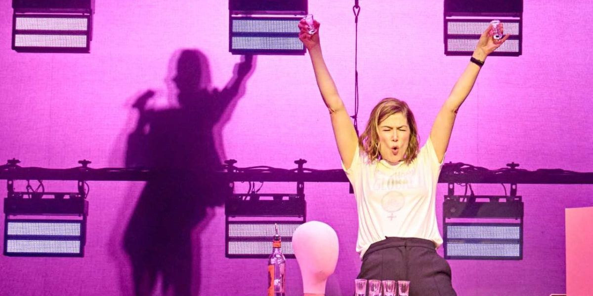A blonde woman holds up a shot glass, with four more lined up in front of her, in Inter Alia on stage