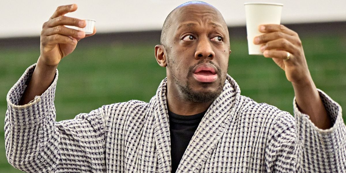 Giles Terera in rehearsals wearing a dressing gown for One Flew Over the Cuckoo's Nest at the Old Vic. He holds two white paper cups up looking concerned.