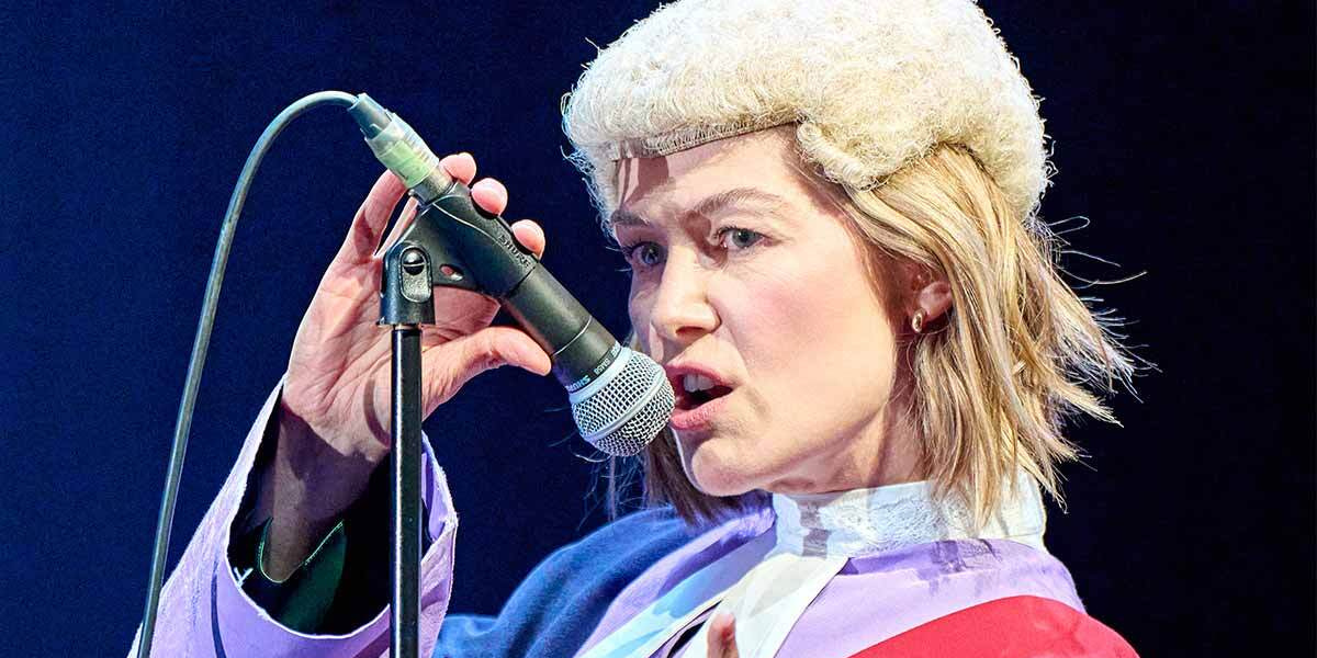 Rosamund Pike wears a classic British judge's wig and looks side on as she holds a microphone