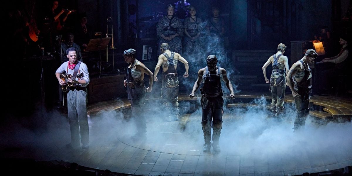 Marley Fenton & the cast of Hadestown. Wearing a red neckerchief and denim overalls playing a guitar and singing. The cast face toward the audience with clenched fists.© Brinkhoff-Moegenburg