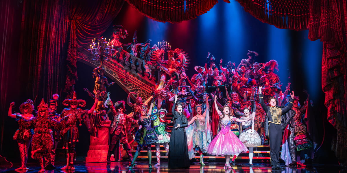 Phantom of the Opera company during Masquerade on a grand staircase in lavish costumes.